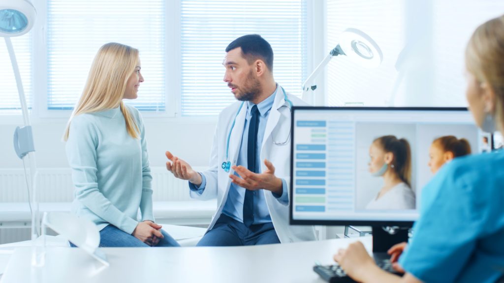 In a cosmetic surgery consultation, ask all the questions you need, including those about the cosmetic surgeon's qualifications and costs for the procedure. In this image, a woman speaks with a plastic surgeon in the background while a depiction of a facelift is shown on a monitor.