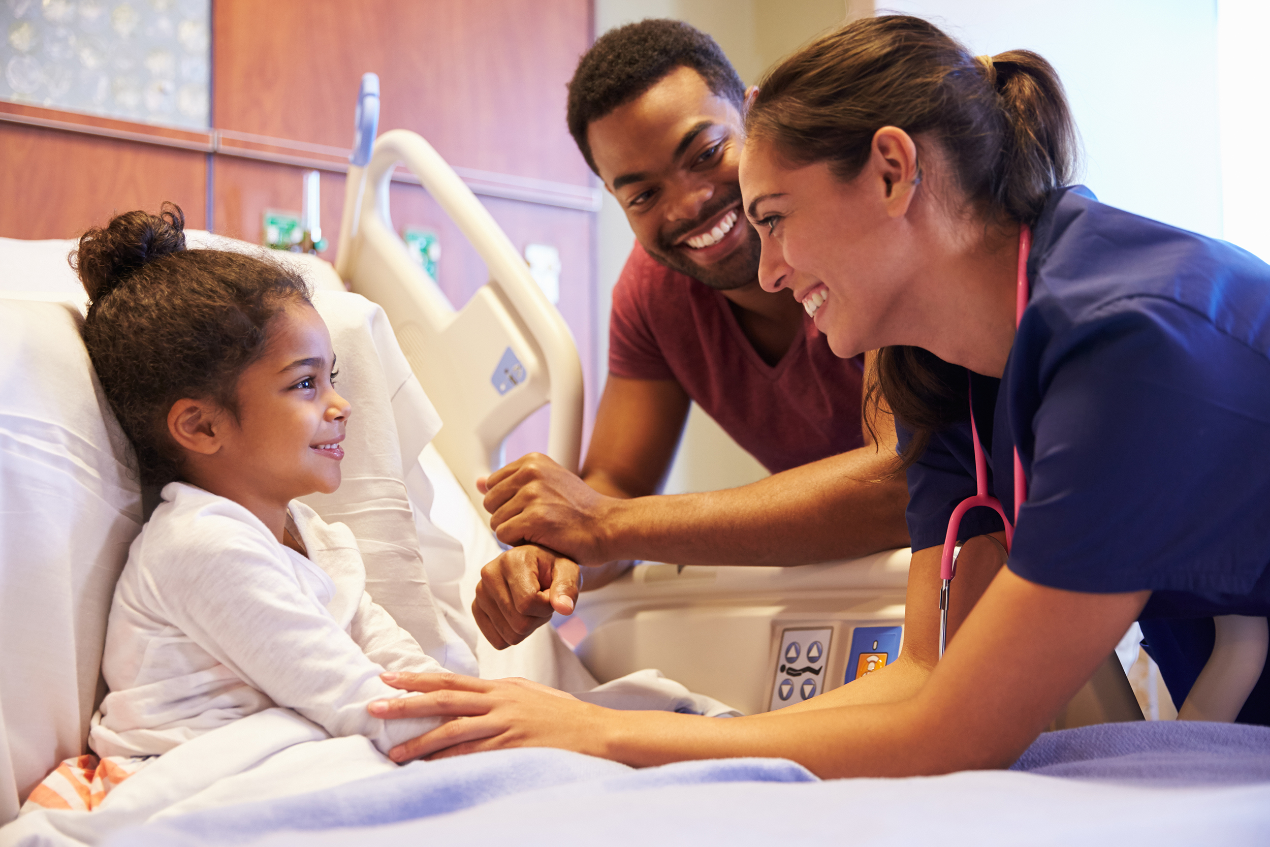 A nurse and a parent talk with a smiling child in a hospital bed in this image, which represents the pediatric surgery process at our Troy, Michigan center.